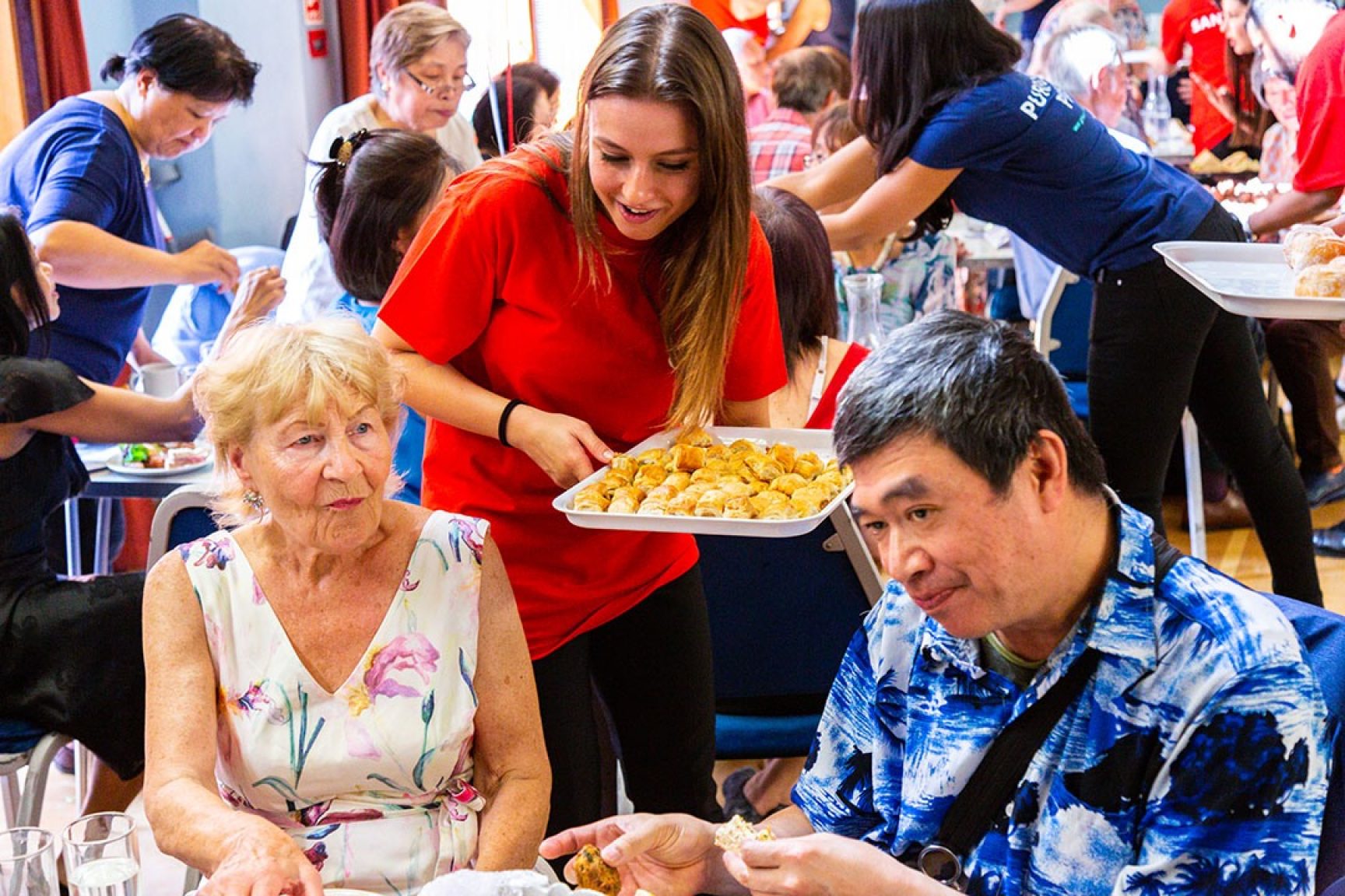 Two people being served at a table by a woman  holding a tray of food.