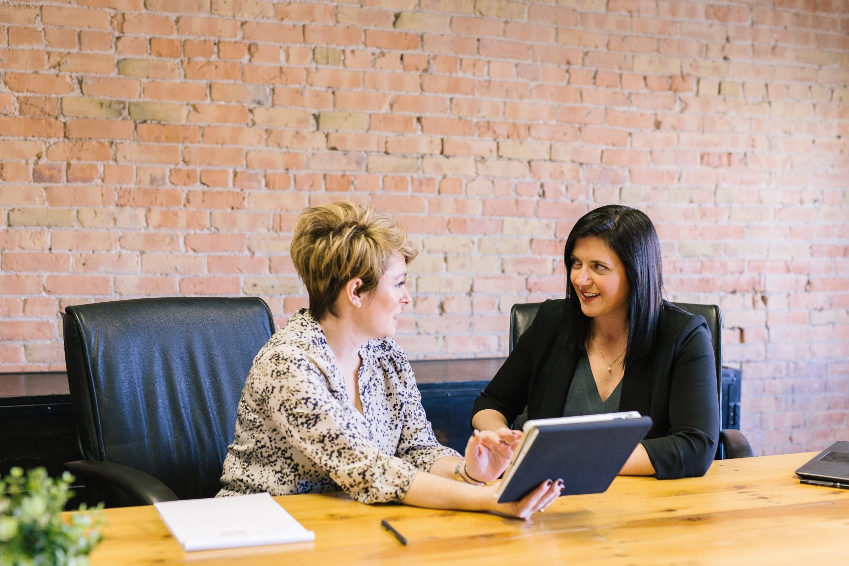 Two women, have a meeting a large table. They are smiling.