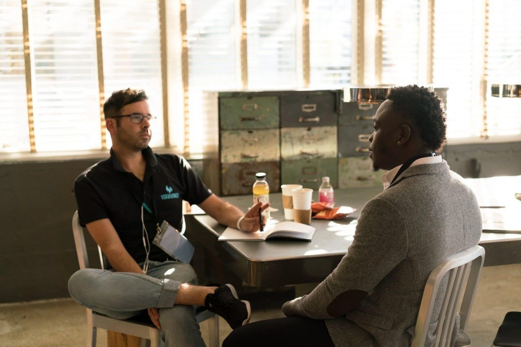 Two men at a table. They are chatting, and one is taking notes.